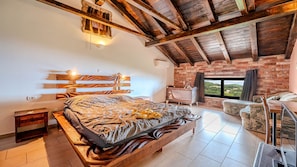 Ein Schlafzimmer mit einem hölzernen Bett, einer Backsteinwand, Blick durch ein Fenster und einer Holzbalkendecke.