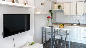 Una cocina moderna con armarios blancos, un televisor negro montado en la pared, una pequeña zona de comedor con mesa y sillas, y una planta en maceta sobre la encimera.