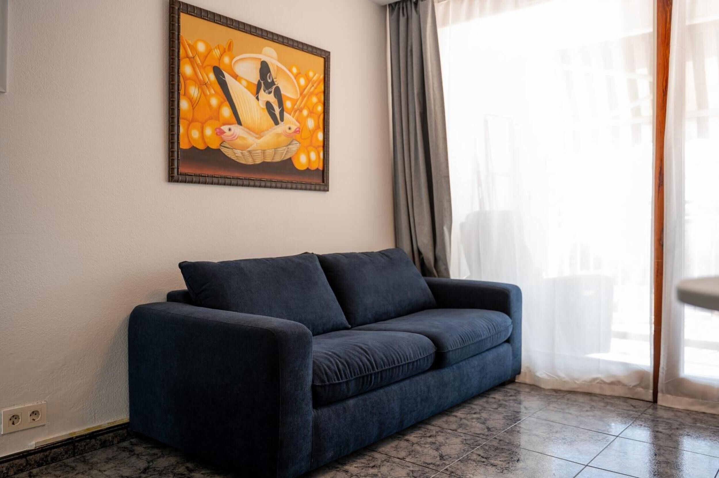 A room with a dark blue sofa, a painting on the wall, and a window with curtains.