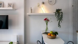 Una cocina moderna con una pared blanca, un reloj, una estantería con objetos decorativos, un florero con flores y una mesa con una cesta de fruta.