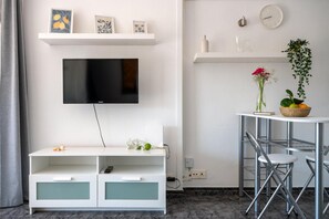 A modern living room with a flat-screen TV, white shelves with decorative items, a round table with chairs, and a small white cabinet with drawers. - Vacation Apartment 'El Mirador' with Sea View, Shared Pool and Wi-Fi (Costa de Adeje)