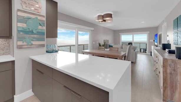 A modern kitchen with a large island, a dining area with a view, and a living space with a large window. - Leonardo Arms 623 Beachfront 2B/2B Vacation Condo! Panoramic Gulf View! (Fort Myers Beach)