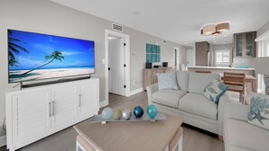 A modern living room with a flat-screen TV, a white sofa, a coffee table with decorative balls, and a kitchen in the background. - Leonardo Arms 623 Beachfront 2B/2B Vacation Condo! Panoramic Gulf View! (Fort Myers Beach)