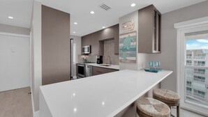 A modern kitchen with a white countertop, stainless steel appliances, and a view of the city through a large window. - Leonardo Arms 623 Beachfront 2B/2B Vacation Condo! Panoramic Gulf View! (Fort Myers Beach)