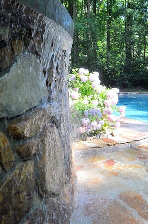 A stone water feature with a cascading waterfall, a swimming pool, and pink flowers in the background. - Skyefall Mountain Retreat: Hot Tub, Fire Pit & Wooded Views | Great for Families (Berkeley Springs)