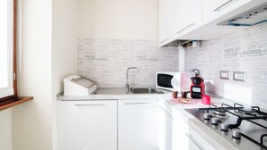 Una cocina compacta con armarios blancos, un lavamanos de acero inoxidable, un microondas, un hervidor rojo y una estufa a gas.