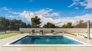 Et rektangulært swimmingpool med klart, blåt vand, omgivet af en flisbelagt terrasse. Tre sorte liggestole står opstillet langs poolens kant.