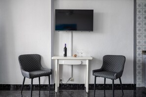 Huoneessa on seinälle asennettu taulutelevisio, valkoinen pöytä, jossa on kaksi tuolia, sekä pullo ja lasit pöydällä.