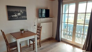Una stanza con un tavolo da pranzo, sedie, una TV su un mobile, un termosifone e un'ampia finestra con tende.