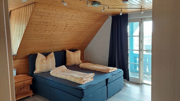 Camera da letto con soffitto spiovente, un letto con copriletto decorato, un comodino e una finestra con tende.