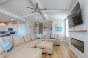 A modern living room with a sectional sofa, a fireplace, a ceiling fan, and a television mounted on the wall. - Experience oceanfront luxury at Tortuga by the Sea in Oak Island, NC. (Oak Island)