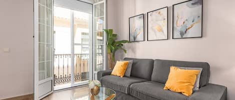 A living room with a grey sofa, a glass coffee table, and a large window with a balcony.