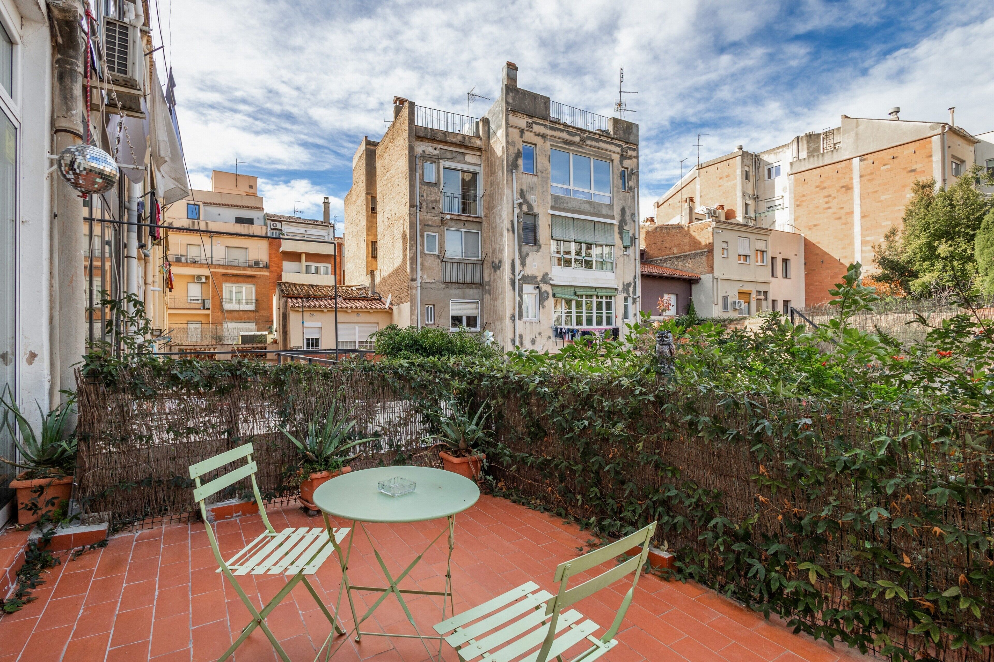 Una terraza con una mesa y sillas, rodeada por un muro alto con plantas, y edificios al fondo.