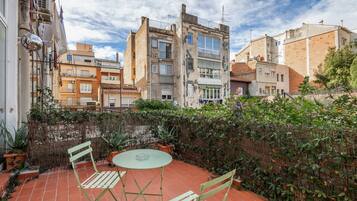 Una terraza con una mesa y sillas, rodeada por un muro alto con plantas, y edificios al fondo.