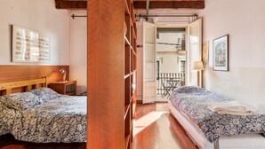 Ein Zimmer mit zwei Betten, Holzboden, Holzbalkendecke, einem kleinen Tisch mit einer Lampe und einer Tür zu einem weiteren Raum.