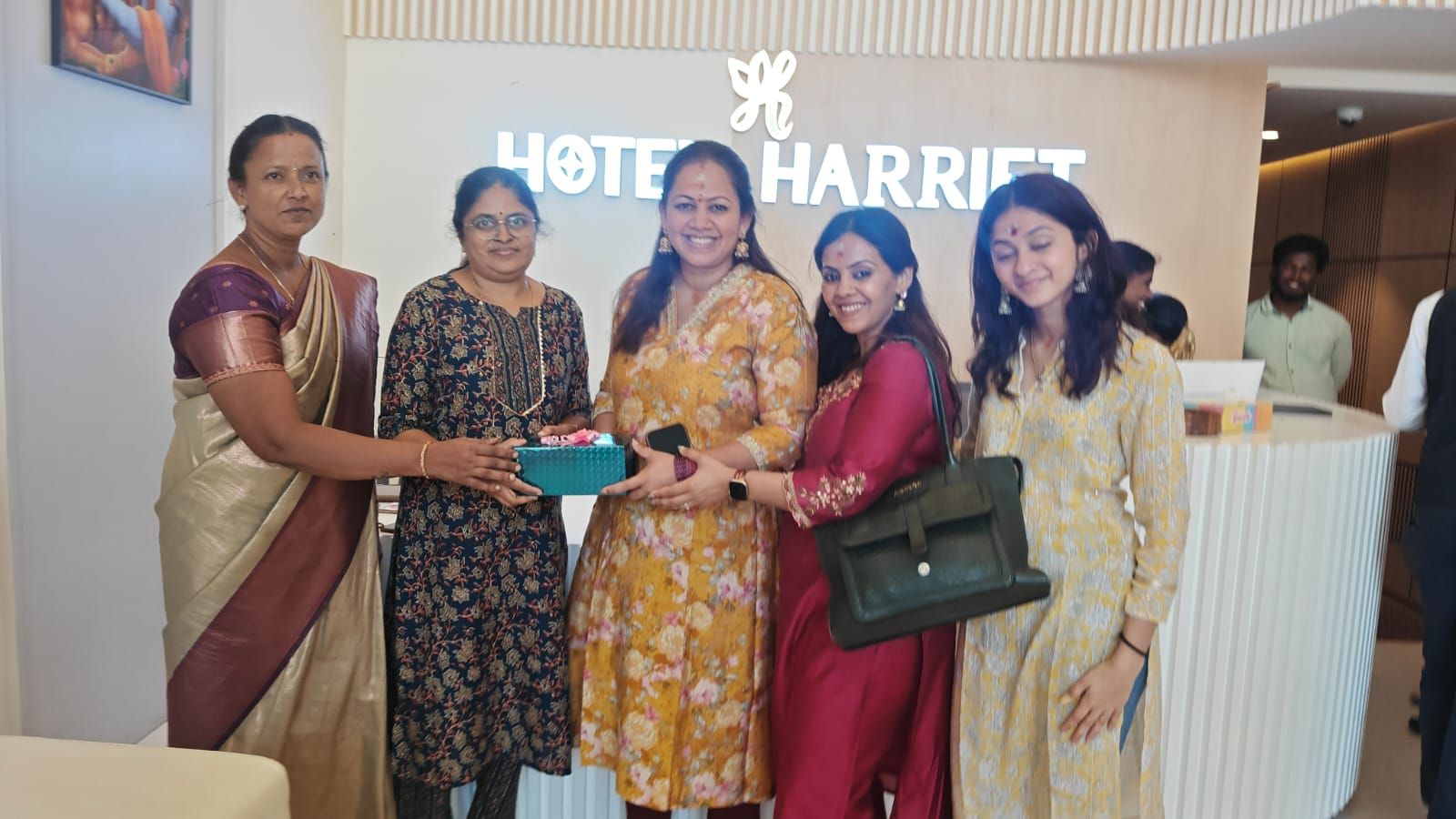 A group of people standing in front of a hotel reception area with a sign that reads 'HOTEL HARRIET'.