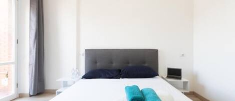 A hotel room with a bed, two pillows, a laptop on a bedside table, and a window with curtains.