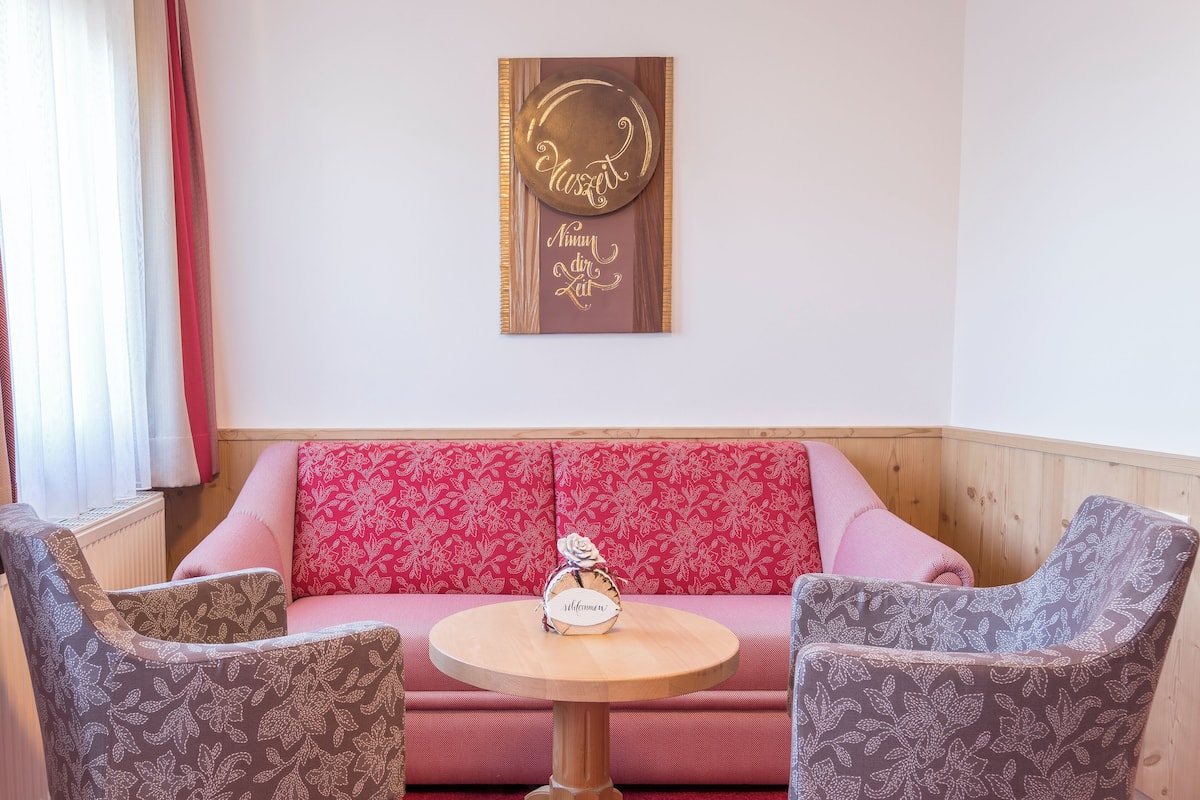 Un rincón acogedor con una mesa redonda de madera, dos sillones estampados y un sofá tapizado rojo. Un objeto decorativo con la inscripción