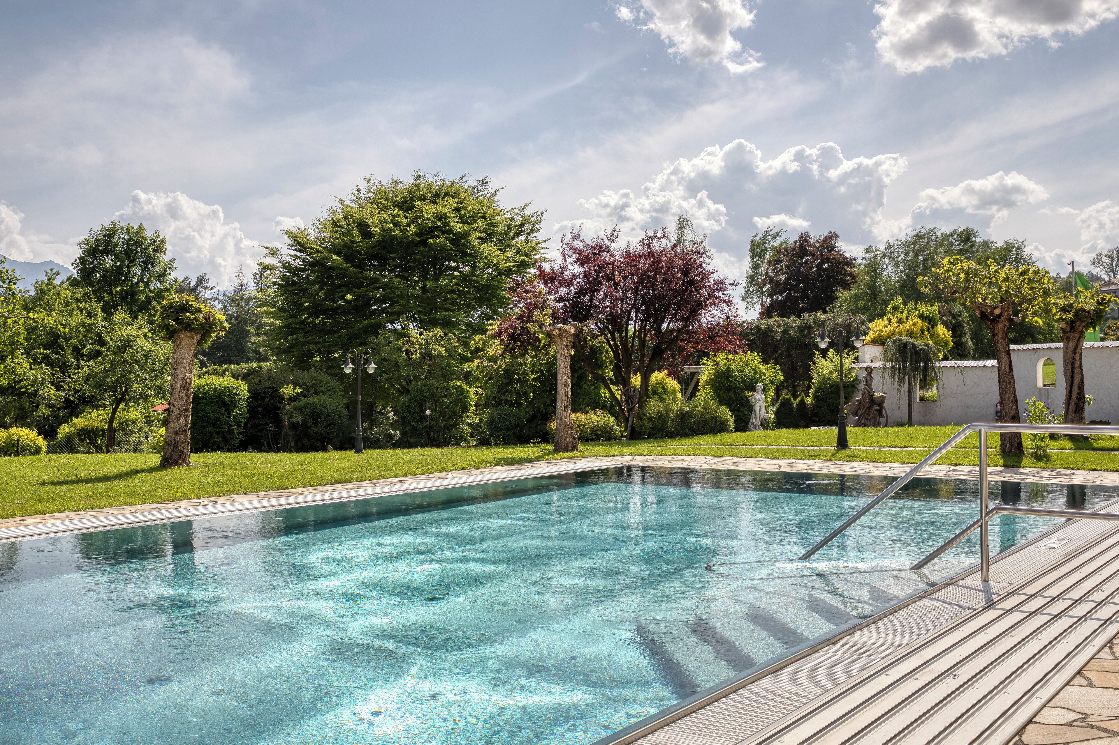 Une piscine aux eaux cristallines, dotée d’une terrasse en bois et de rampes en métal, entourée d’une végétation luxuriante et d’arbres.