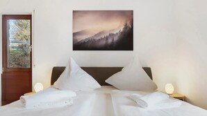 A neatly made bed with white linens and pillows, a painting of a forest on the wall, and a door leading to a balcony. - Lotti Apartment with Mountain Views, Balcony, and Wi-Fi (Titisee-Neustadt)