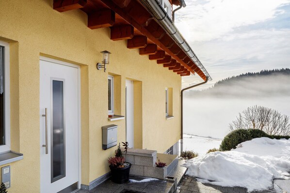 A building with a yellow facade, a white door, a small porch with steps, and a view of a snowy landscape and forest. - Holiday Apartment Sonnenstüble with Mountain View, Shared Terrace, and Wi-Fi (Titisee-Neustadt)