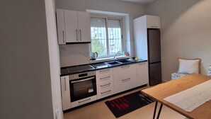 A modern kitchen with white cabinets, a black countertop, a built-in oven, a refrigerator, and a window with blinds. - AR Hotel (Tirana)