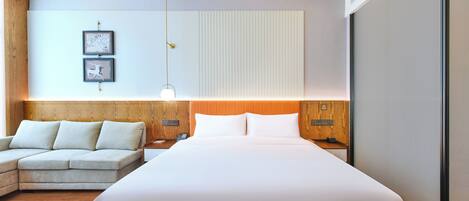 A modern hotel room with a large bed, a sofa, a wooden headboard, and a wall-mounted light fixture.