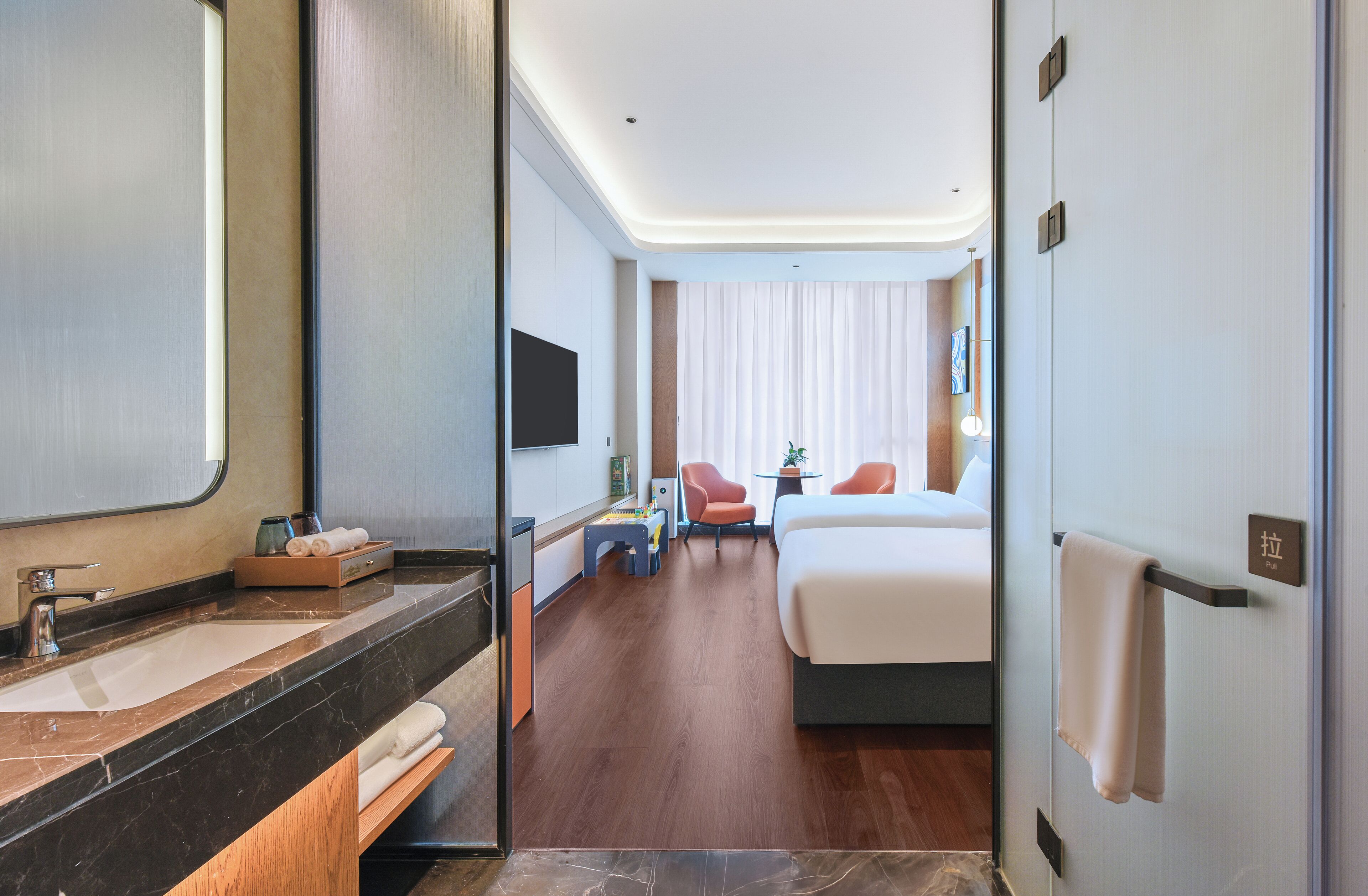 Een hotelkamer met een groot bed, een bureau met stoel, een kleine tafel met een plant en een badkamer met spiegel en wastafel.