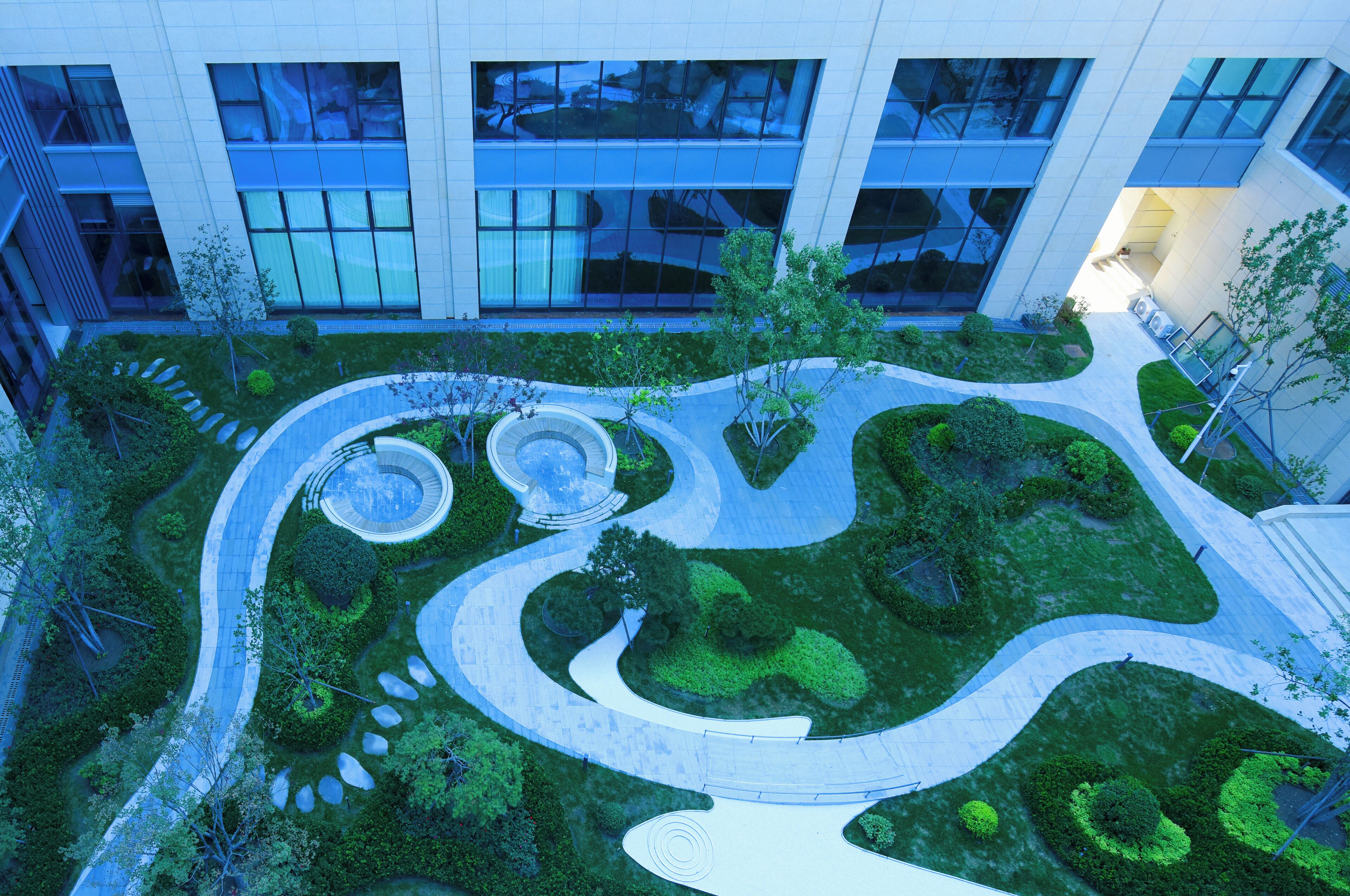 An aerial view of a landscaped courtyard with winding pathways, circular planters, and a variety of trees and shrubs.