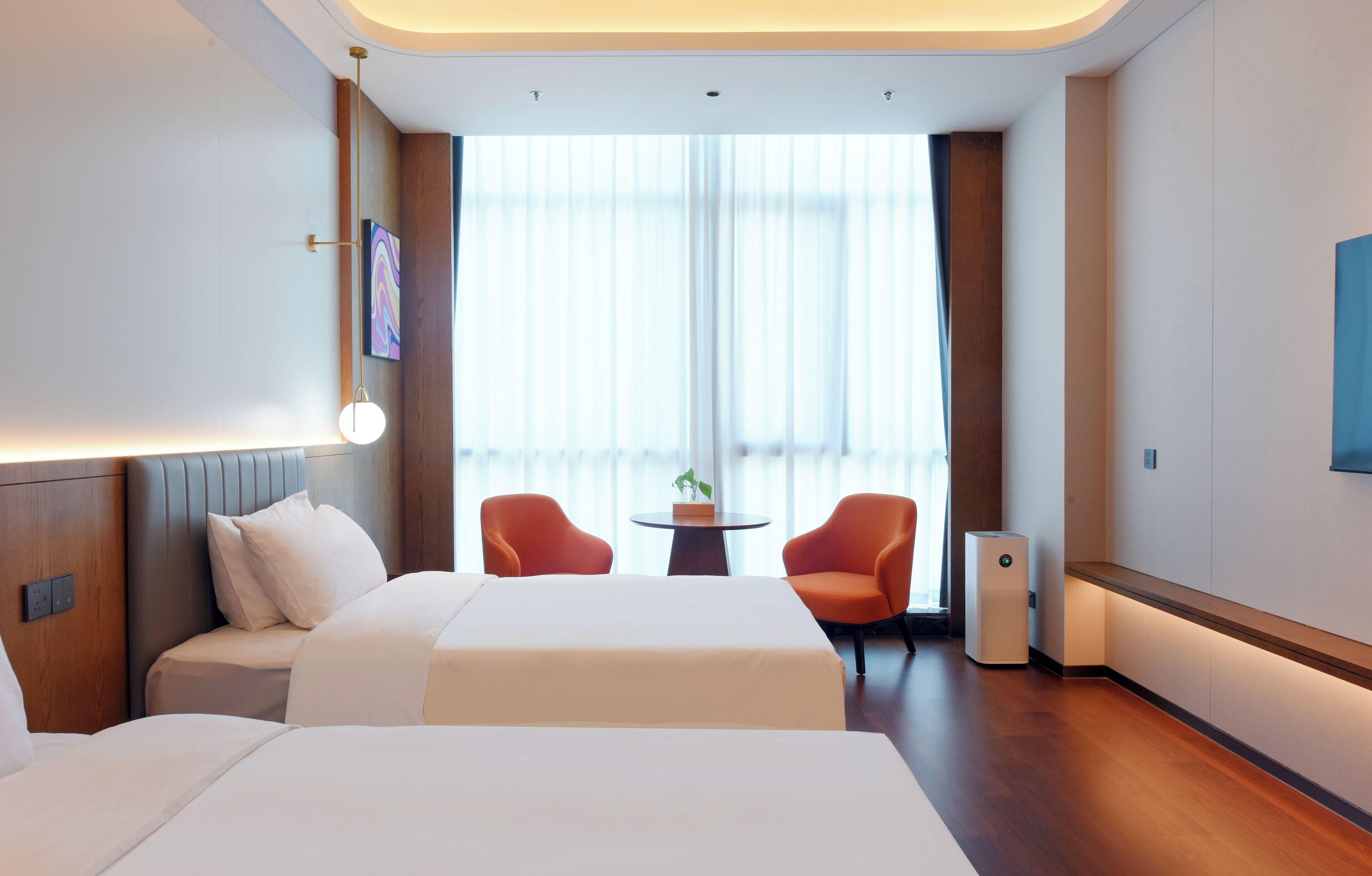 A hotel room with two beds, a small table, two orange chairs, a potted plant, and a large window with curtains.