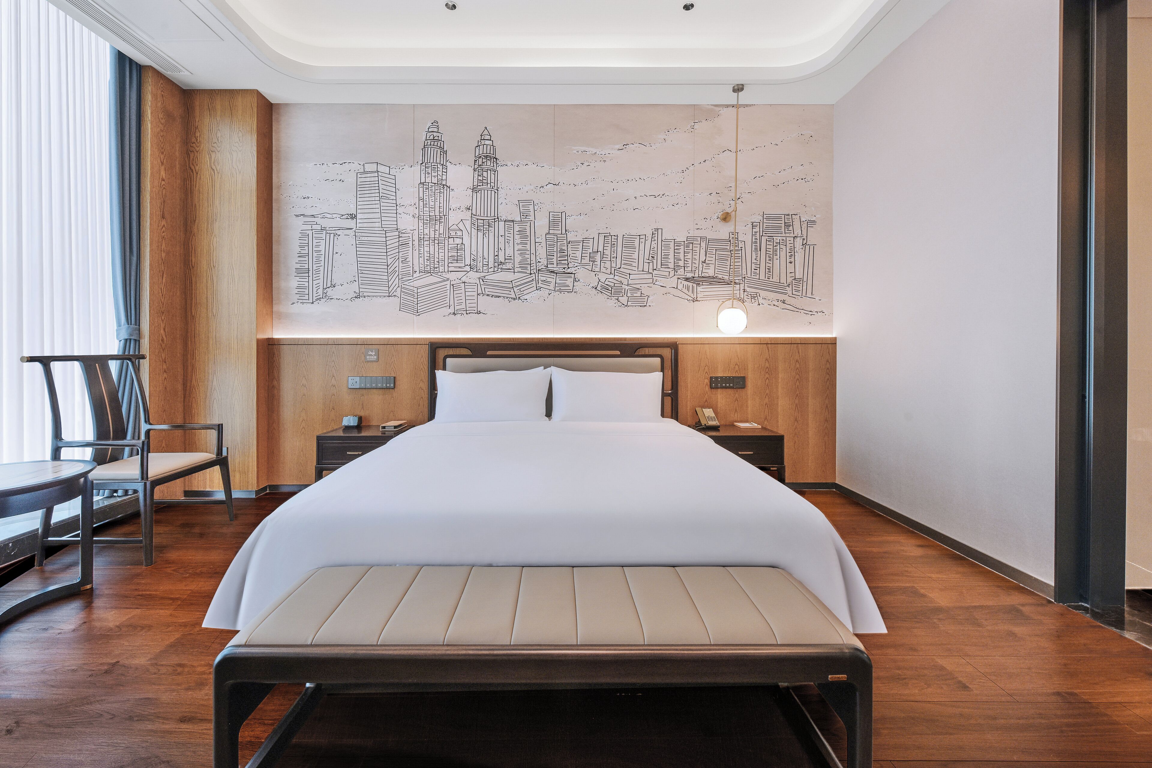 Una habitación de hotel moderna con una cama grande, cabecero de madera, un mural de paisaje urbano en la pared, un escritorio con una silla y una mesita de noche con una lámpara.