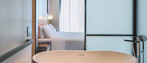 A modern bathroom with a freestanding bathtub, a vanity with a mirror, and a bedside table with a lamp.