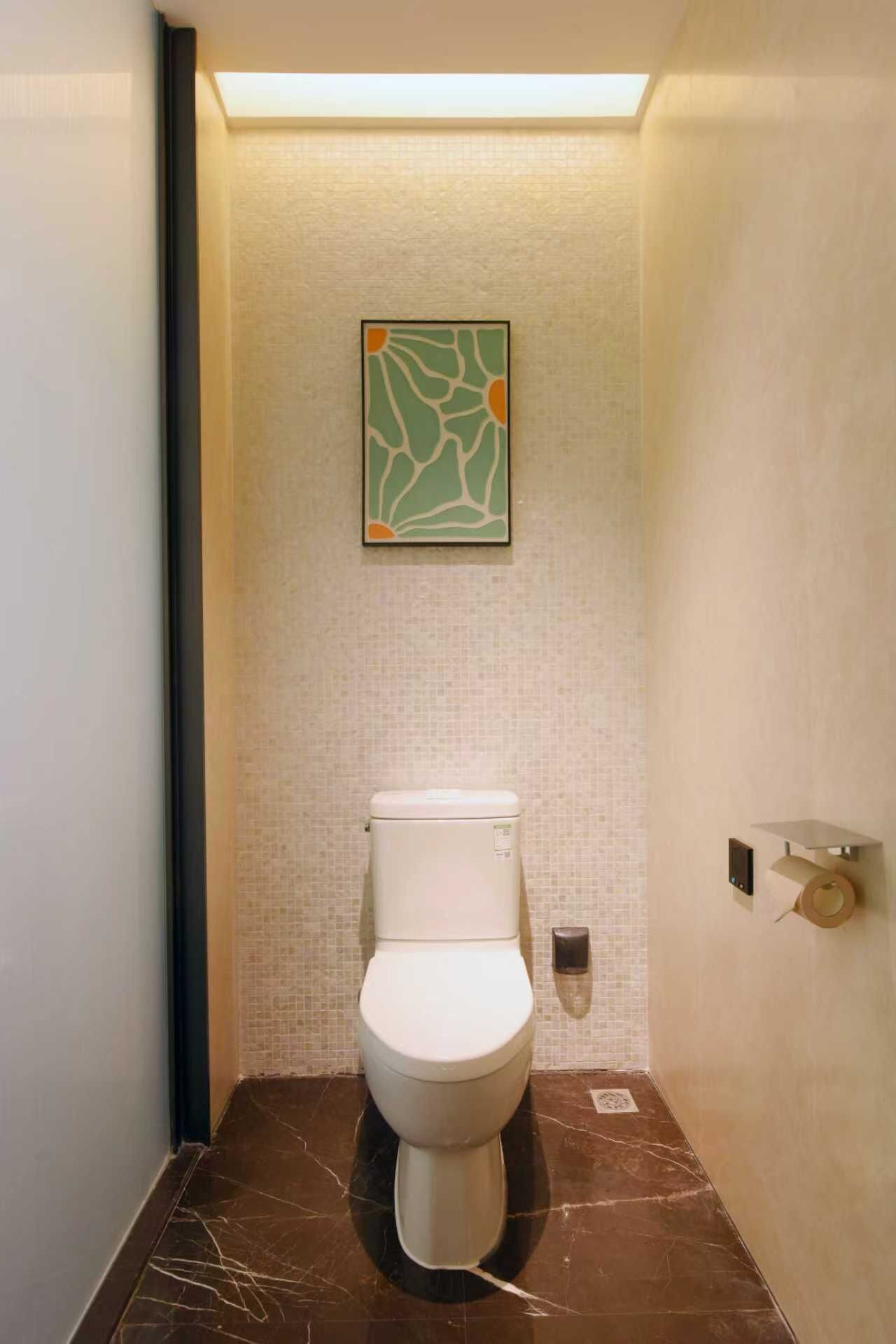 Et moderne badeværelse med toilet, toiletpapirholder på væggen og et indrammet abstrakt kunstværk over toilettet.