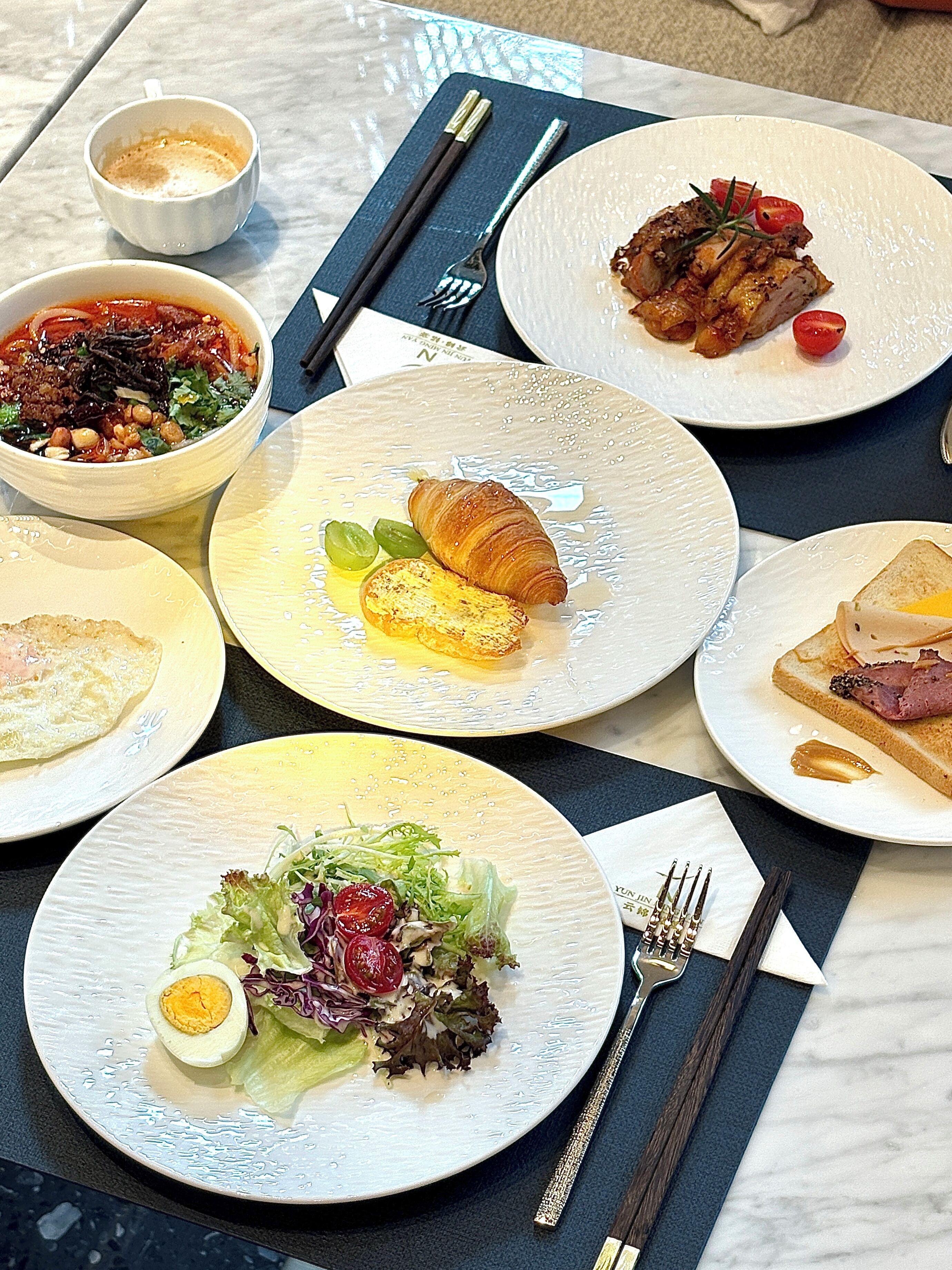 Ein Tisch reichhaltig gedeckt mit verschiedenen Speisen, darunter ein Croissant, Salat und Fleisch, sowie Essstäbchen und einer Serviette auf einer Marmortafel.
