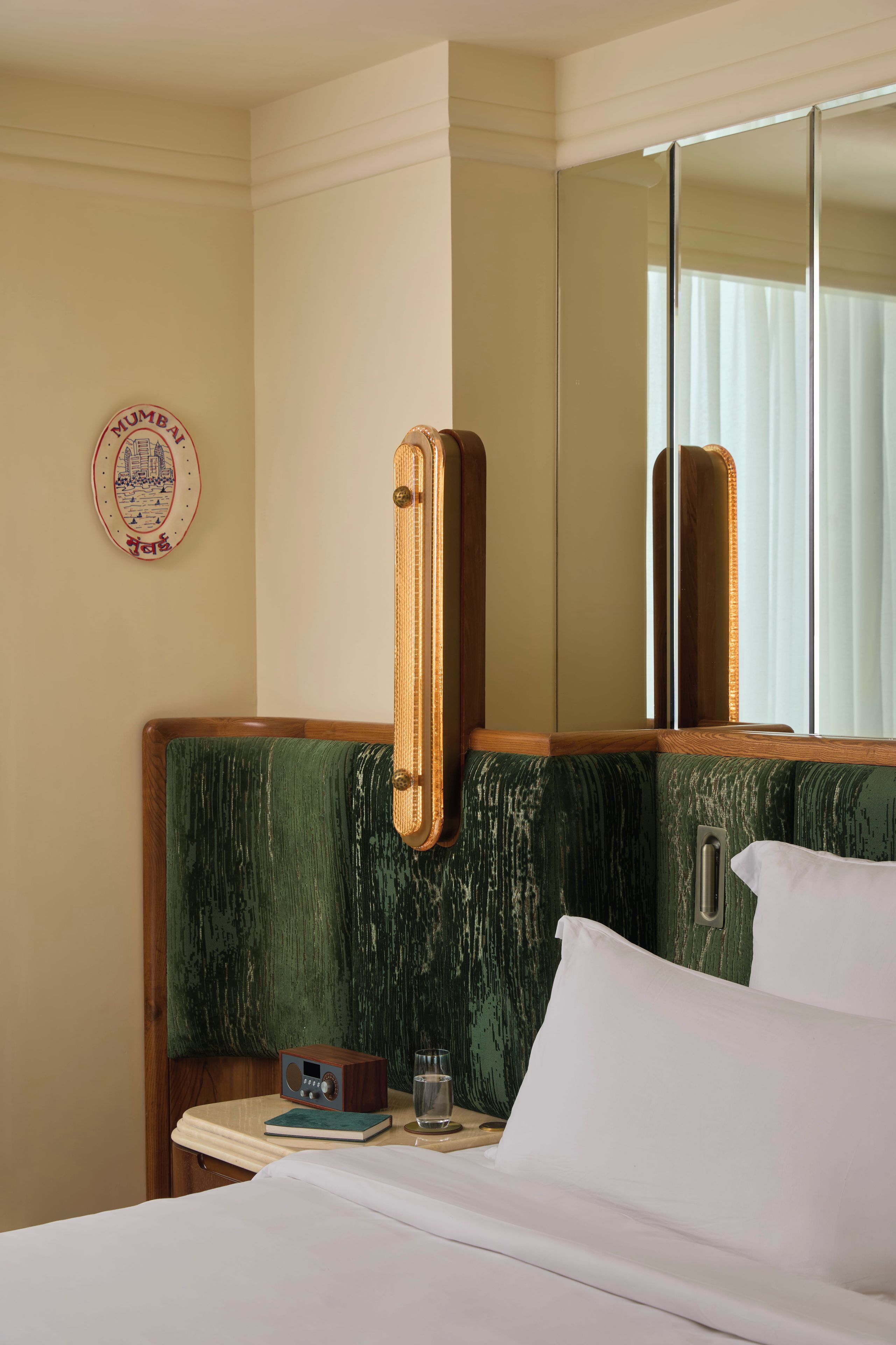 Habitación de hotel con una cama, una mesita de noche con un libro y un vaso, un reloj de pared y un espejo.