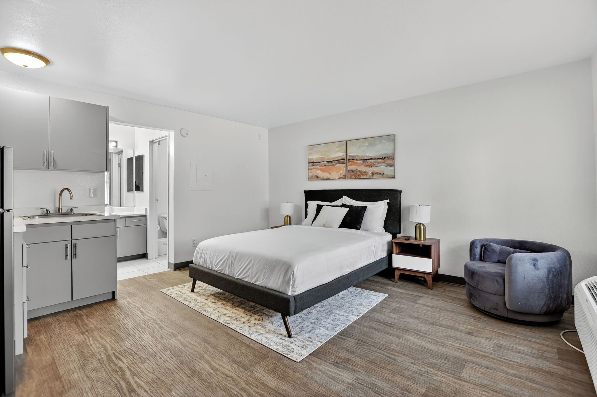 A modern bedroom with a bed, bedside tables, a chair, and a view of the kitchen.