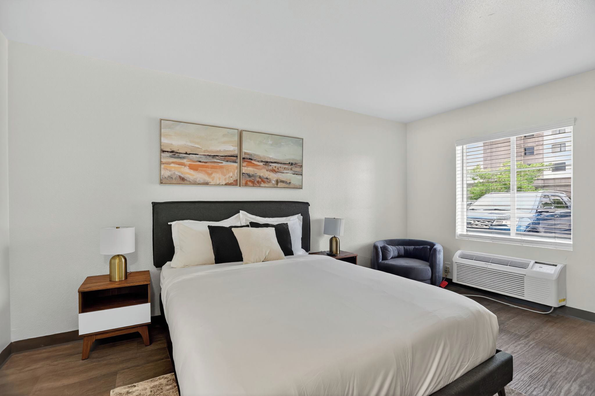 Una habitación de hotel moderna con una cama grande, dos mesitas de noche con lámparas, una silla y una ventana con persianas.