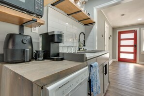 Une cuisine équipée d’une machine à café, d’une bouilloire et d’un lave-vaisselle. On y trouve une étagère en bois au-dessus du plan de travail et une porte rouge à l’arrière-plan.