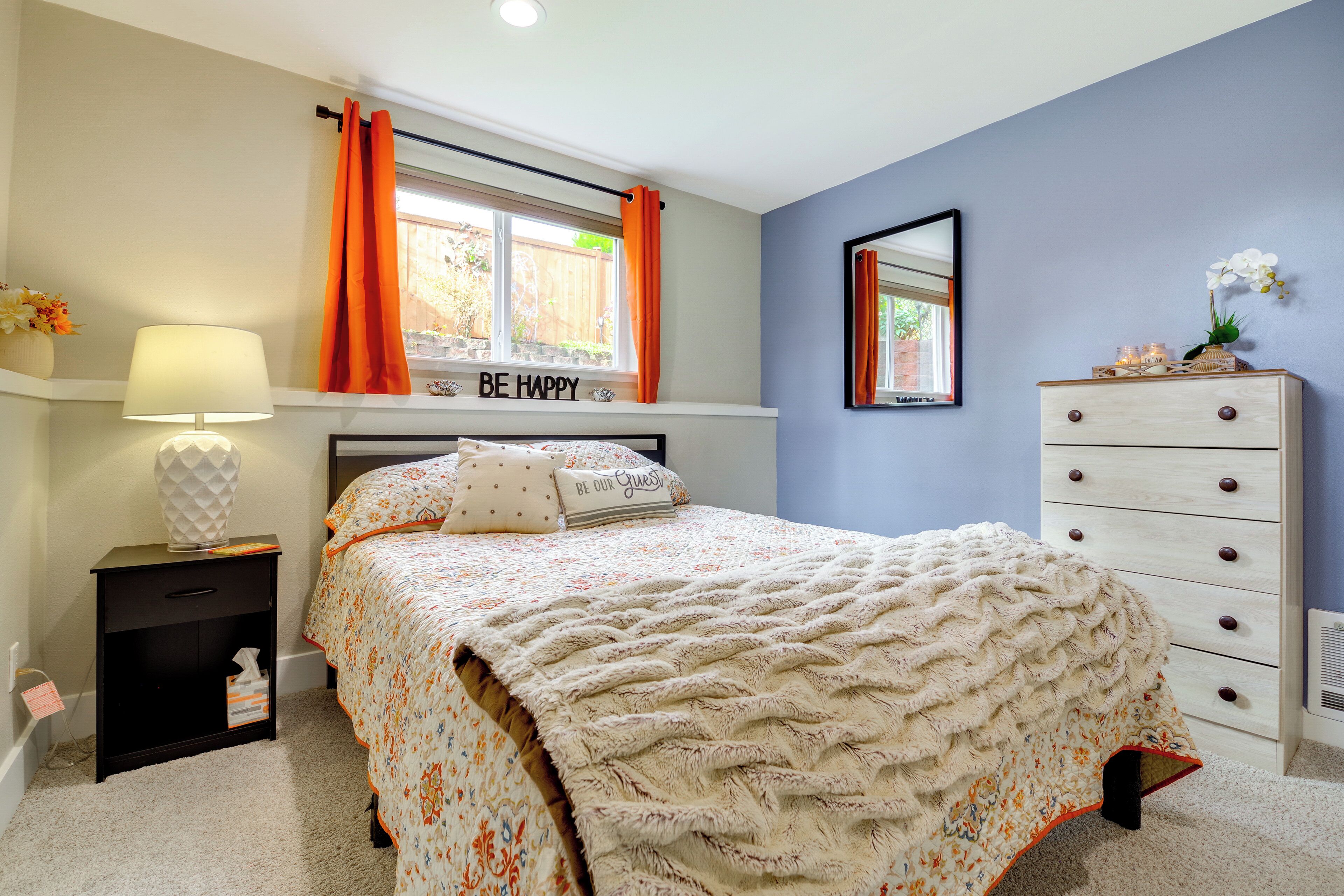 Une chambre à coucher avec un lit, une table de chevet, une commode, une fenêtre avec des rideaux orange et un miroir au mur.