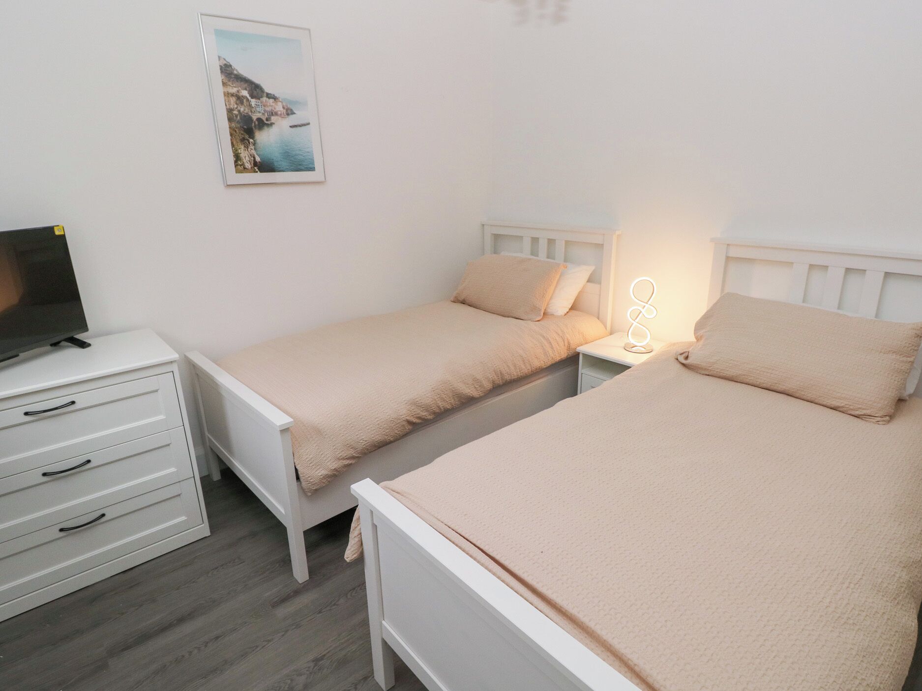 A bedroom with two single beds, a bedside table with a lamp, a TV on a stand, and a framed picture on the wall.