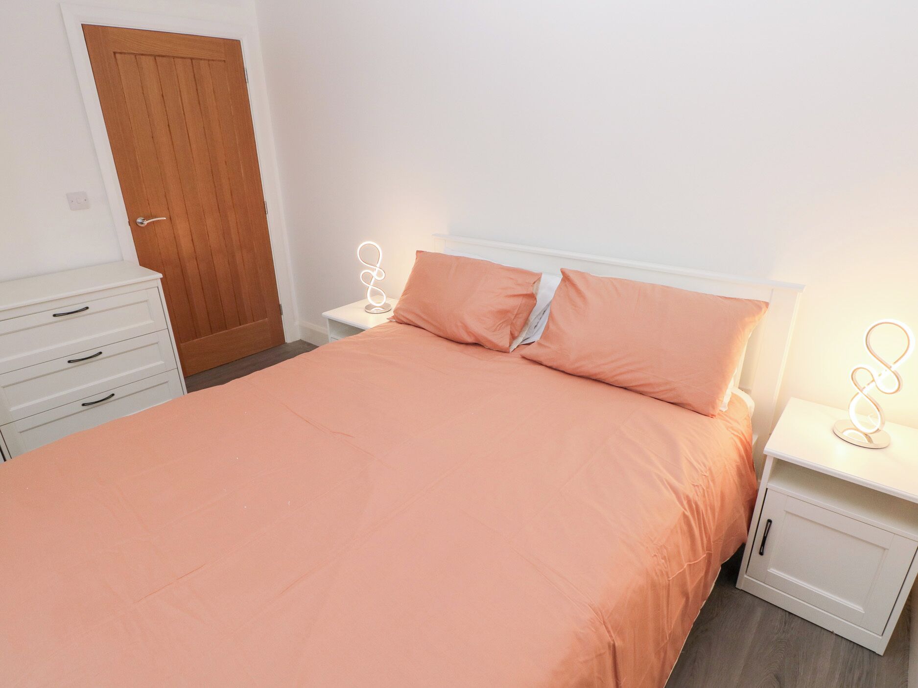 Quarto com cama, mesas de cabeceira brancas, porta de madeira e cômoda branca.