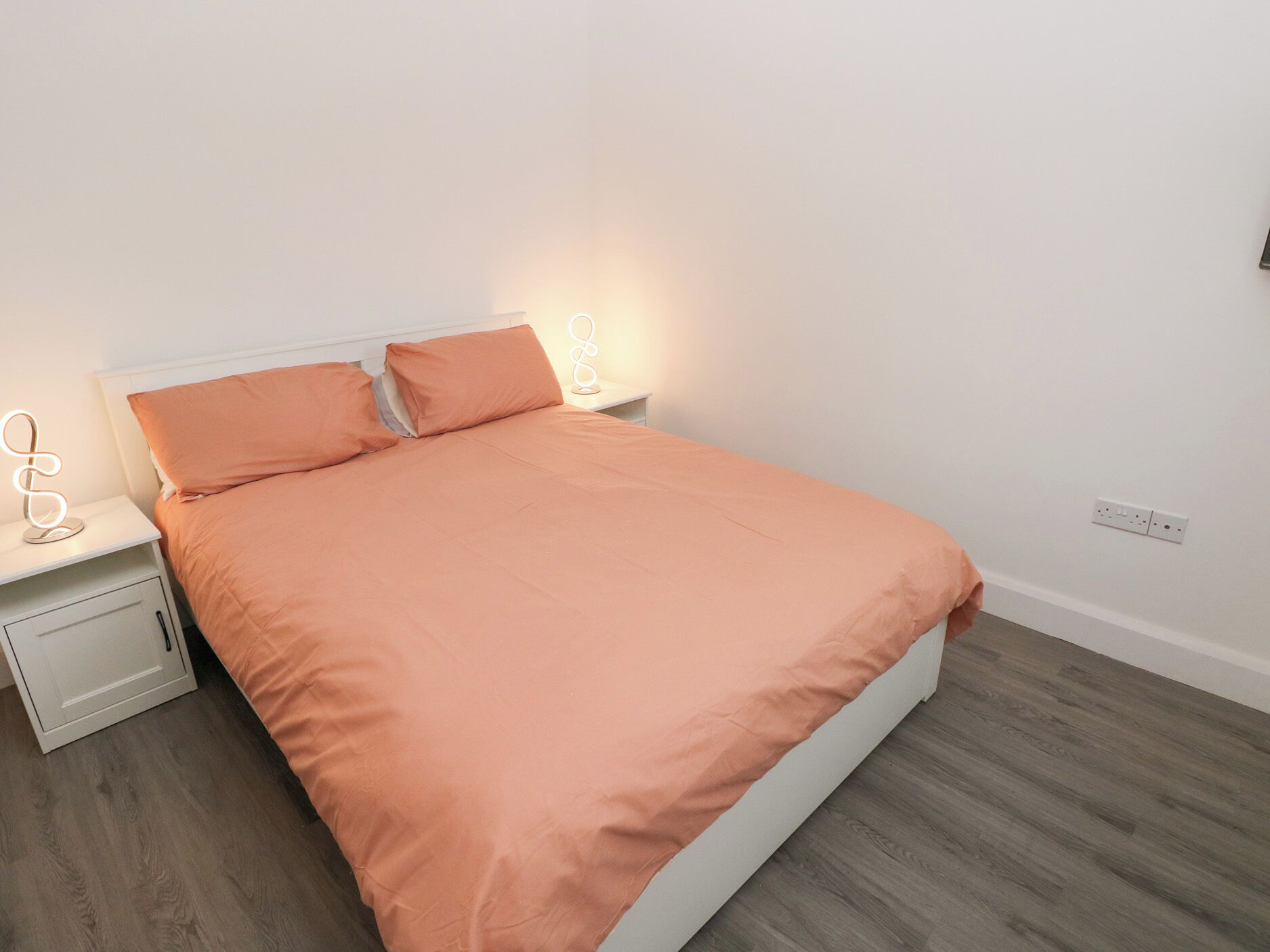 Una cama bien hecha con una colcha color melocotón y almohadas a juego, flanqueada por mesitas de noche blancas con una lámpara moderna.