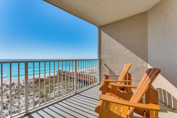 A balcony with a view of the beach and ocean, wooden chairs, and a railing.