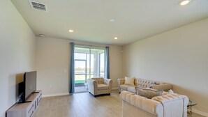 Un salon spacieux avec un téléviseur à écran plat posé sur un meuble en bois, deux fauteuils beiges, un canapé beige et une table basse en verre.
