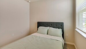 Une chambre avec un lit simple, une tête de lit rembourrée de couleur grise et une fenêtre avec des stores.