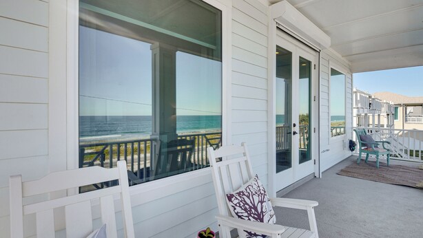 A covered outdoor area with white wooden walls and large windows overlooking the ocean. There are white chairs and a small table with a plant. - Oceanfront Retreat in Flagler Beach! Bright Apt (Flagler Beach)
