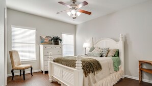 Une chambre à coucher avec un lit blanc, un ventilateur de plafond, une commode, une chaise et une fenêtre avec des stores.