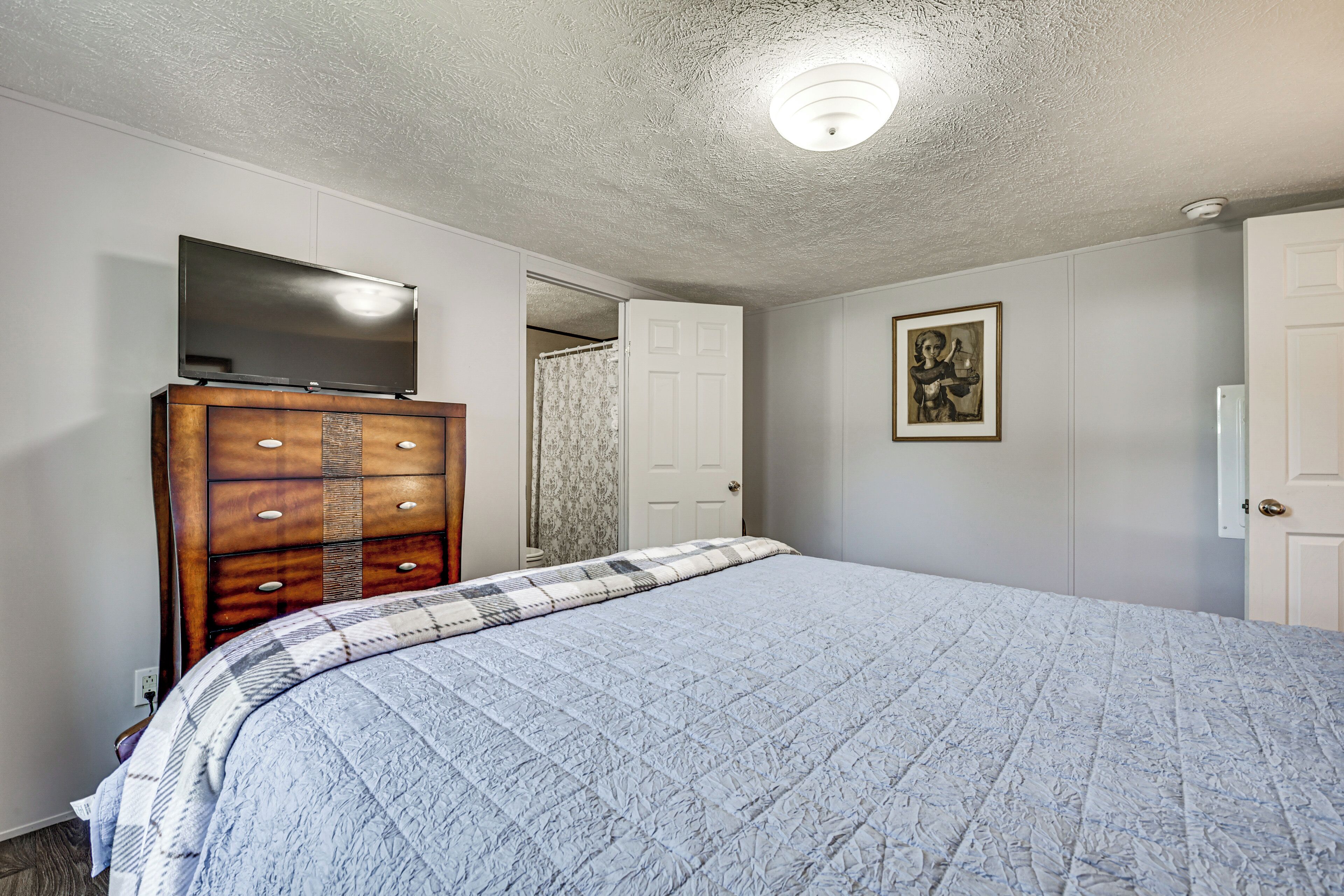 A bedroom with a large bed, a wooden dresser, a flat-screen TV, and a door leading to another room.