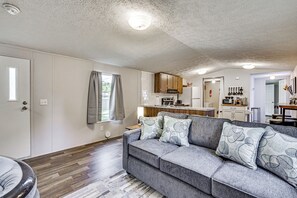 A modern living room with a grey sofa, wooden flooring, and a kitchen in the background.