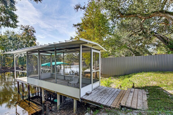 A covered deck with a glass enclosure, a wooden dock, and a grassy area with a fence.